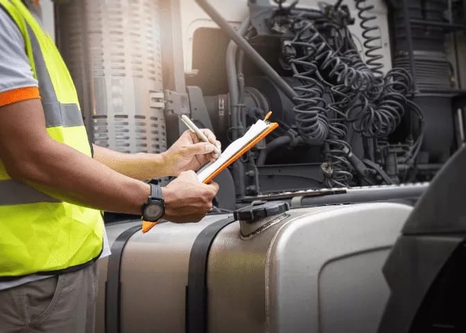 Heavy Vehicle engineer in high-visibility vest writing on a clipboard in front of a truck engine with visible cables and mechanical components.